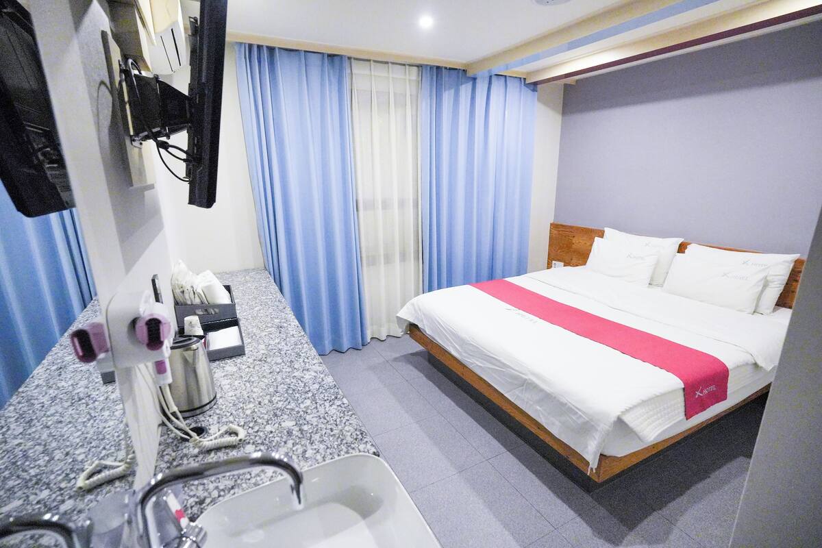 A hotel room with a bed, a sink, a mirror, a TV mounted on the wall, and a window with blue curtains.