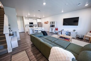Un salon moderne avec un canapé d’angle, un téléviseur à écran plat et une cuisine en arrière-plan.