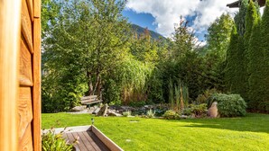 Una terraza de madera que lleva a un jardín exuberante con un estanque, rodeado de diversas plantas y árboles.