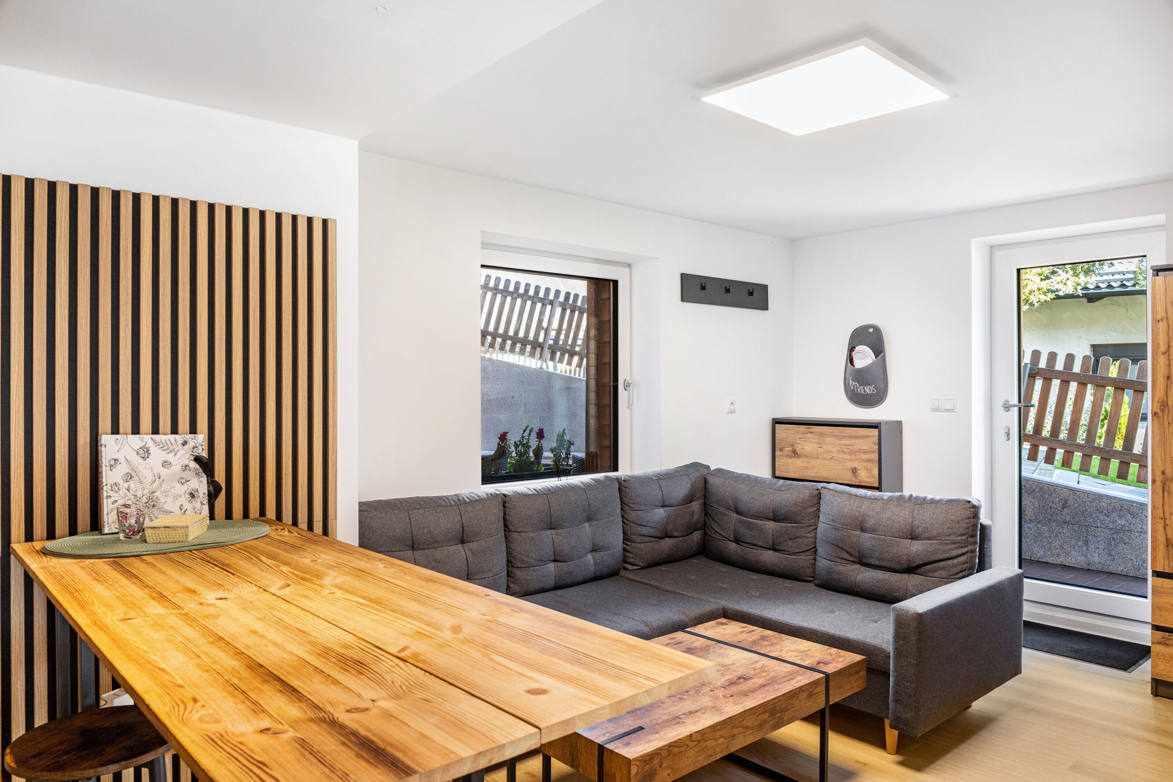 A modern living room with a wooden dining table, grey sectional sofa, and a view of a garden through a door.