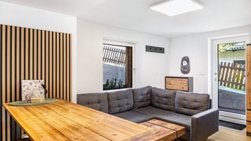 A modern living room with a wooden dining table, grey sectional sofa, and a view of a garden through a door.