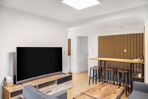 Un salon moderne doté d’un téléviseur à écran plat, d’une table basse en bois, de canapés gris et d’un coin bar avec des tabourets.