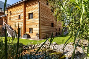 Une cabane en bois avec un chemin de pierres menant à l’entrée, un petit étang parsemé de nénuphars et une cour couverte d’herbe.