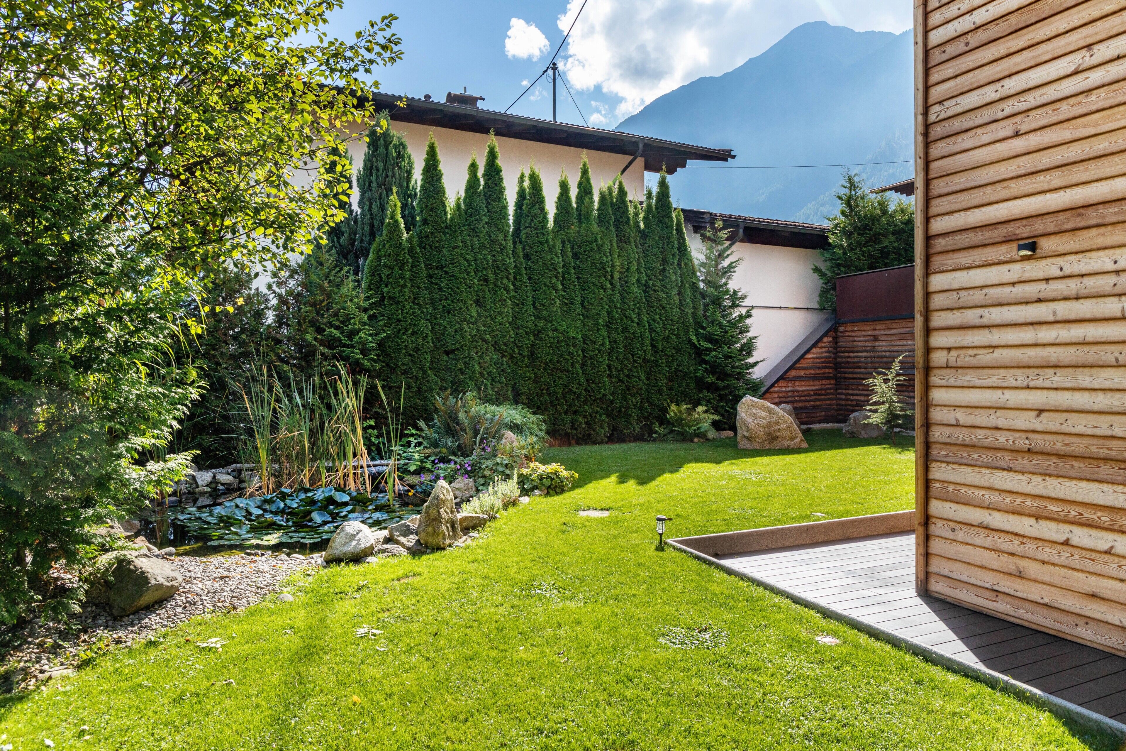 A well-maintained garden with a wooden deck, a stone wall, and a mountain backdrop.