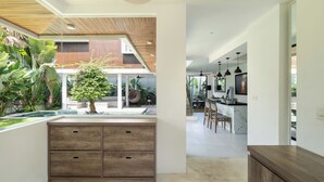 A modern kitchen with a central island, wooden cabinetry, and a view of the outdoor garden. - Melasti Villa 2 by Alfred in Bali (Canggu)