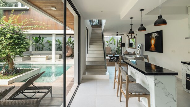 A modern kitchen with a marble countertop, a dining area with chairs, and a view of an outdoor pool area. - Melasti Villa 2 by Alfred in Bali (Canggu)