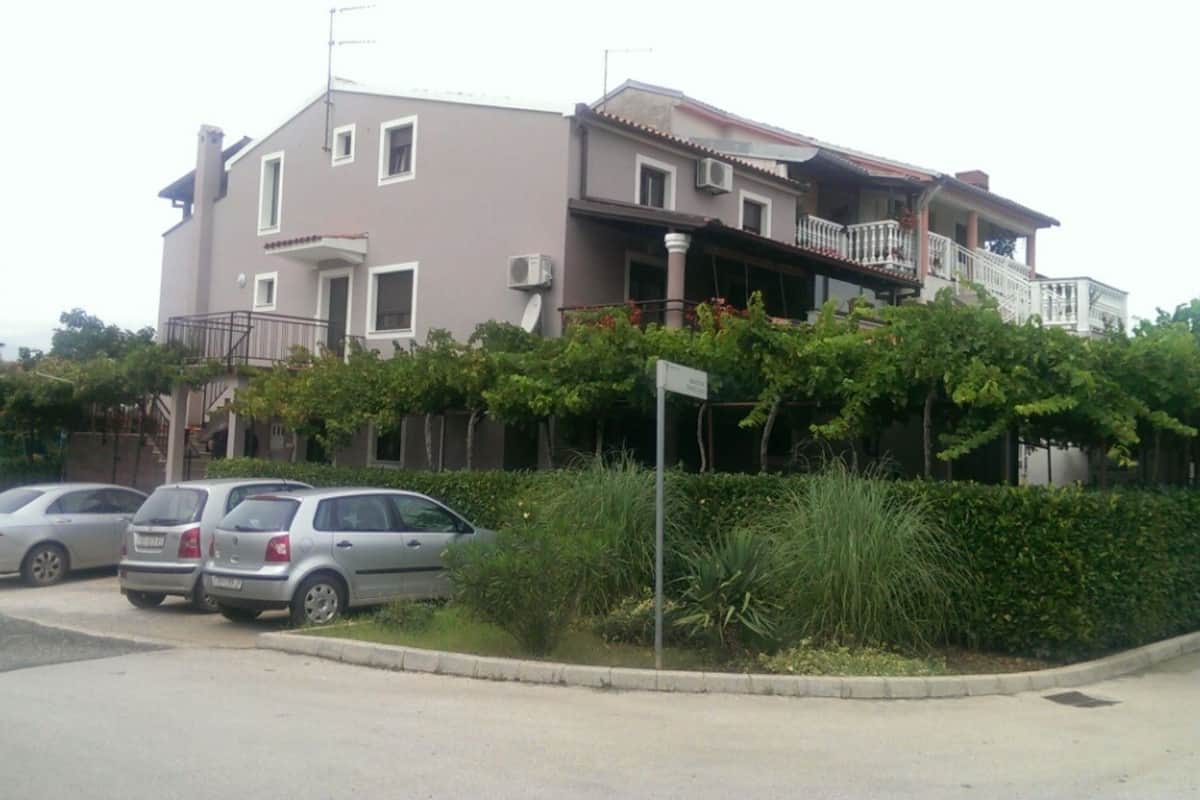 A two-story building with a balcony, parked cars, and greenery in front.