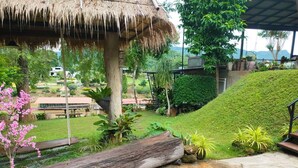 A lush garden with a thatched roof structure, a grassy slope, and various plants. - Rimwang The River Life (Sai Yok)