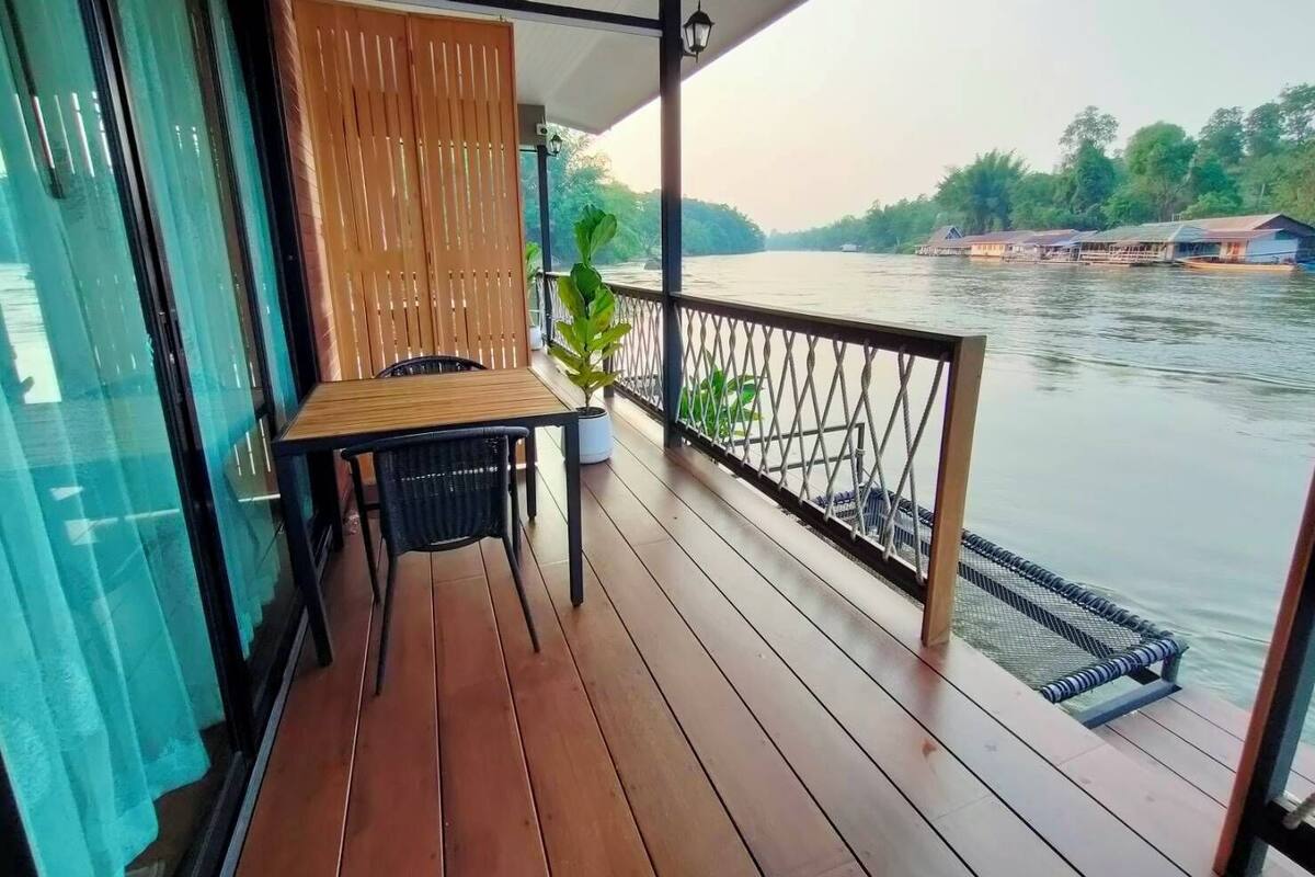 A wooden deck with a table and chairs, overlooking a calm river with boats and greenery in the background.