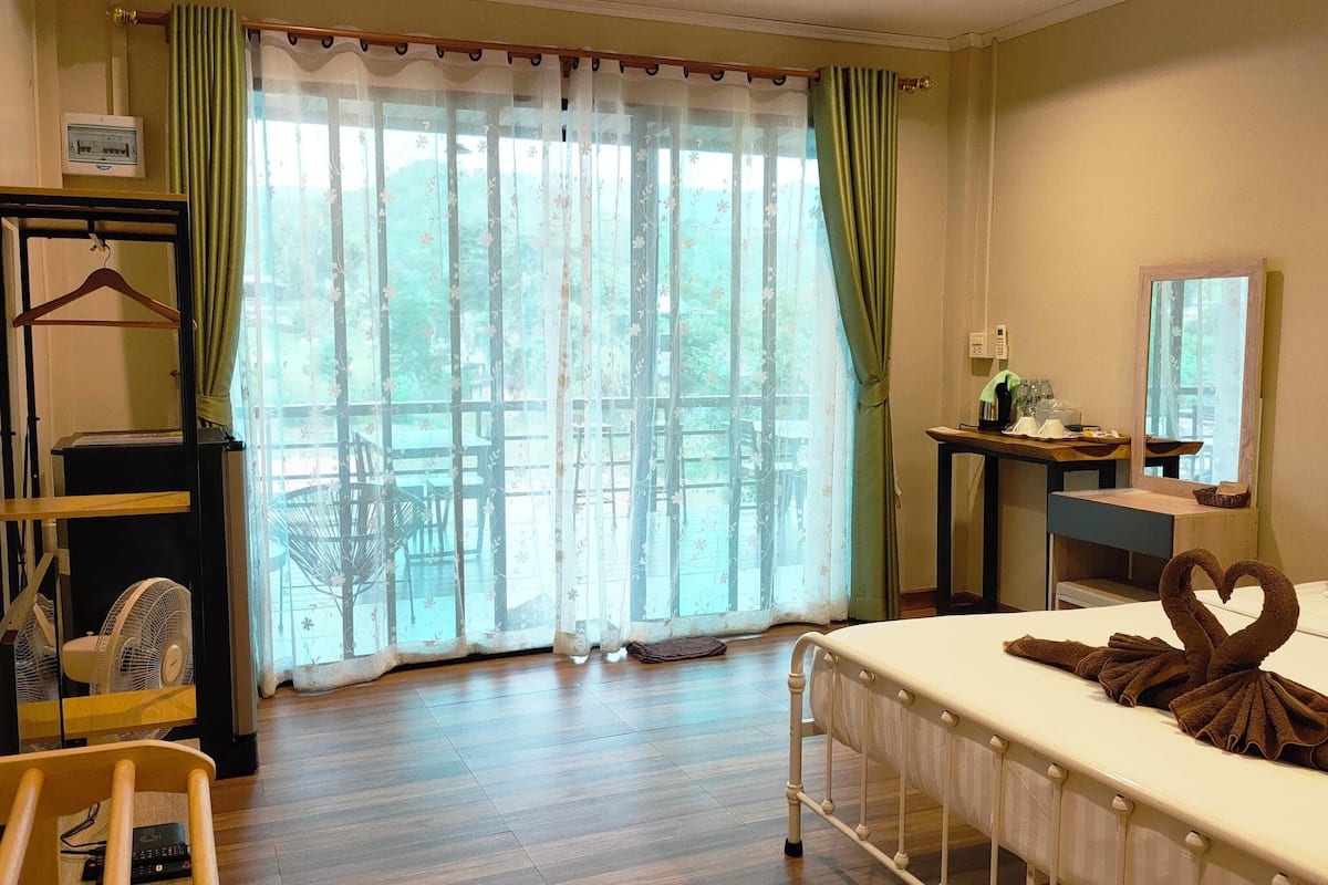 A hotel room with a bed, a desk, a chair, a fan, and a view through a window with curtains.