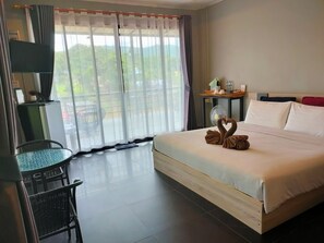 A hotel room with a bed, a small table, a chair, a fan, and a view of greenery outside. - Rimwang The River Life (Sai Yok)