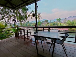 A covered outdoor area with a table and chairs, overlooking a river and distant buildings. - Rimwang The River Life (Sai Yok)