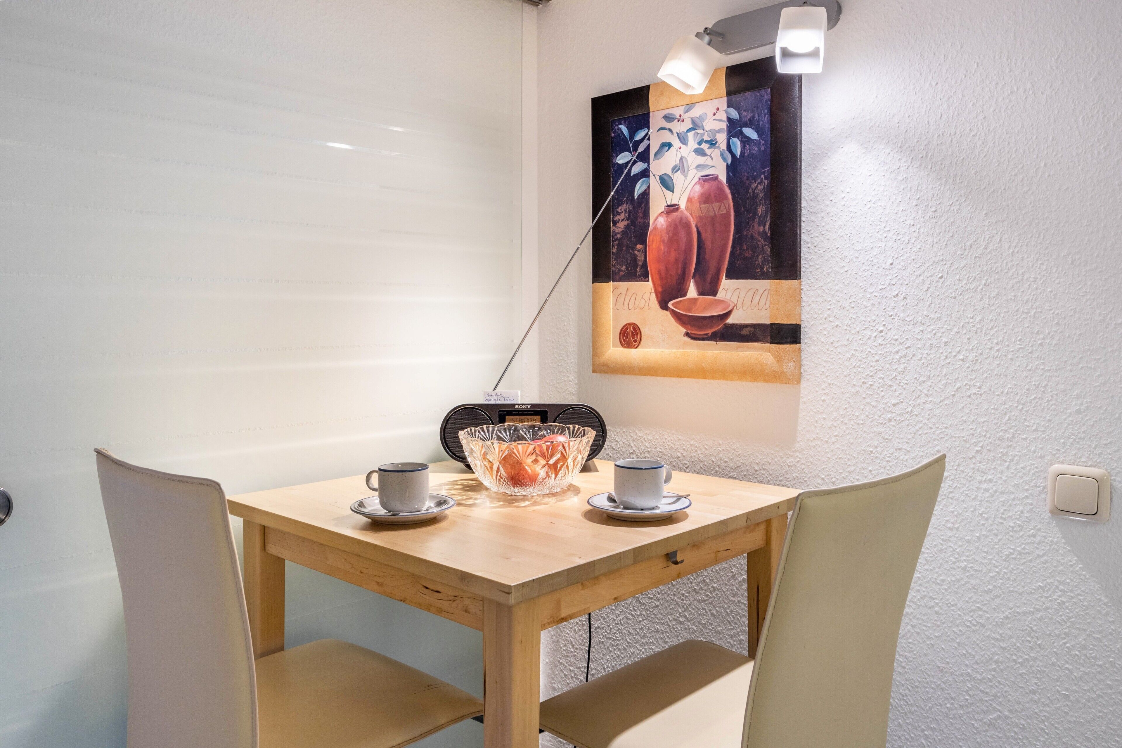 Ein Essbereich mit einem Holztisch, zwei Stühlen, einer Glasvase, zwei Tassen und einer Wandleuchte.