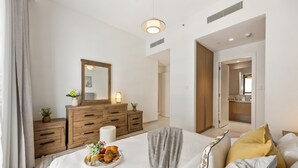 A modern bedroom with a large bed, wooden dresser, and a mirror. - WYZ RENT|Beachside 2BR W Pool Gym Lagoon Access (Dubai)