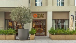 A building with a 'BABA ROMA' sign, potted plants, and a 'WYZRENT' logo. - WYZ RENT|Beachside 2BR W Pool Gym Lagoon Access (Dubai)