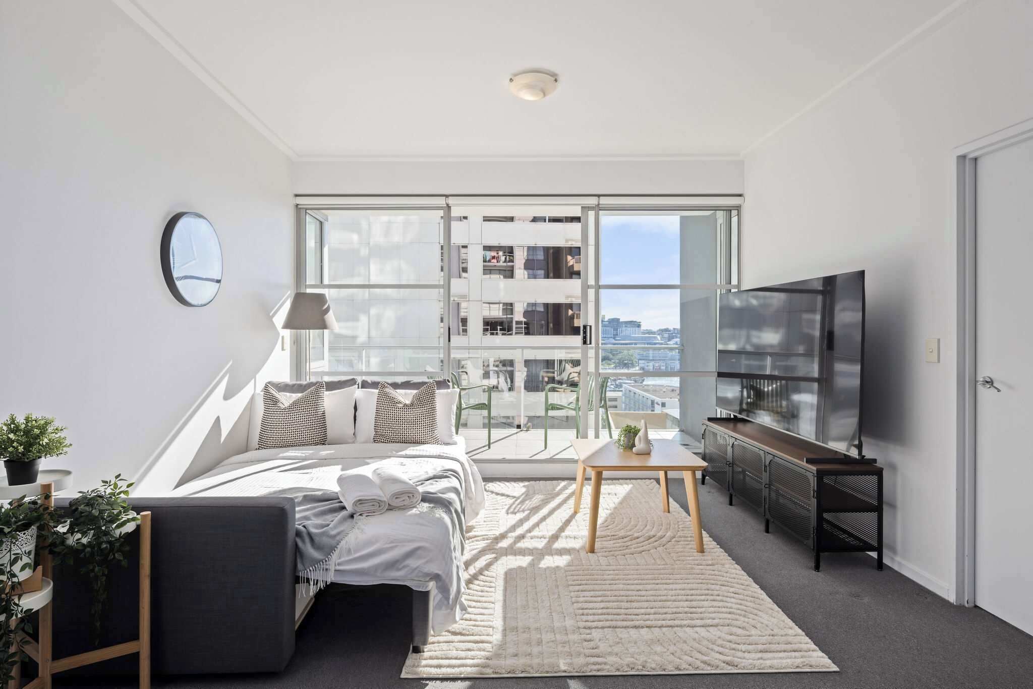 A modern bedroom with a large bed, a TV, and a view of a cityscape through floor-to-ceiling windows.