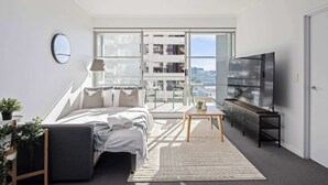 Une chambre moderne avec un grand lit, une télévision et une vue sur le paysage urbain grâce à de grandes fenêtres du sol au plafond.