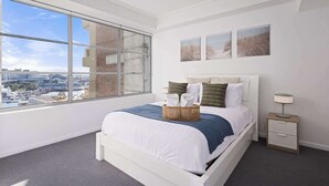 Une chambre moderne avec une grande fenêtre, un lit recouvert de linge de lit blanc, une table de chevet et trois tableaux accrochés au mur.