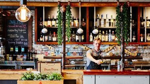 Un bar avec un comptoir en bois, un tableau noir servant de menu et une variété de bouteilles d’alcool.
