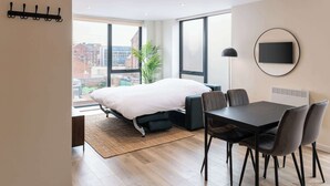 A modern hotel room with a large bed, a dining table with chairs, a TV mounted on the wall, and a view of the city through large windows. - Modern 1BR Central Liverpool Apt with WiFi (Merseyside)
