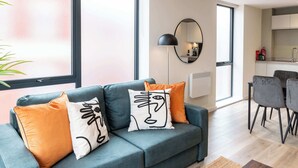 A modern living room with a blue sofa, orange and white cushions, a round mirror, and a dining area with grey chairs. - Modern 1BR Central Liverpool Apt with WiFi (Merseyside)