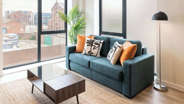 A modern living room with a blue sofa, a glass coffee table, and a large window offering a city view. - Modern 1BR Central Liverpool Apt with WiFi (Merseyside)