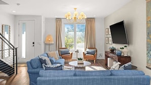 A living room with a blue sofa set, two armchairs, a coffee table, a chandelier, a TV, and a staircase. - 2 Homes-Sleeps 24 | Downtown Great for Big Groups! (Nashville)