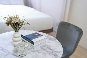 Una habitación ordenada con una cama blanca, una mesa redonda de mármol con un jarrón de flores secas, un libro y un vaso transparente.