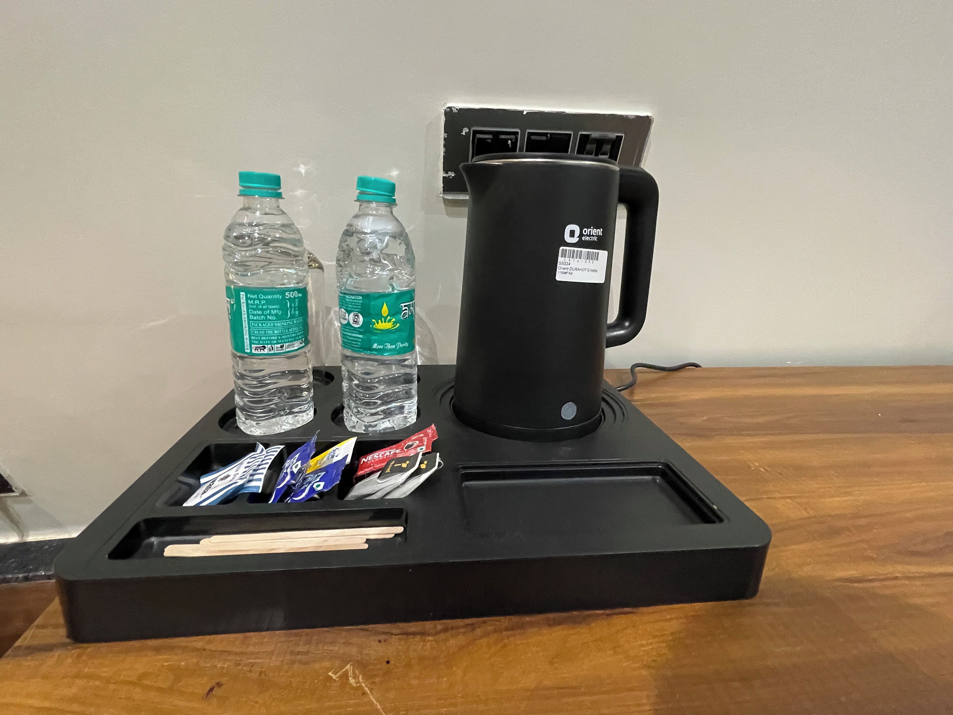 Un plateau noir sur lequel se trouvent deux bouteilles d’eau, une machine à café et plusieurs paquets de collations, le tout posé sur une table en bois.