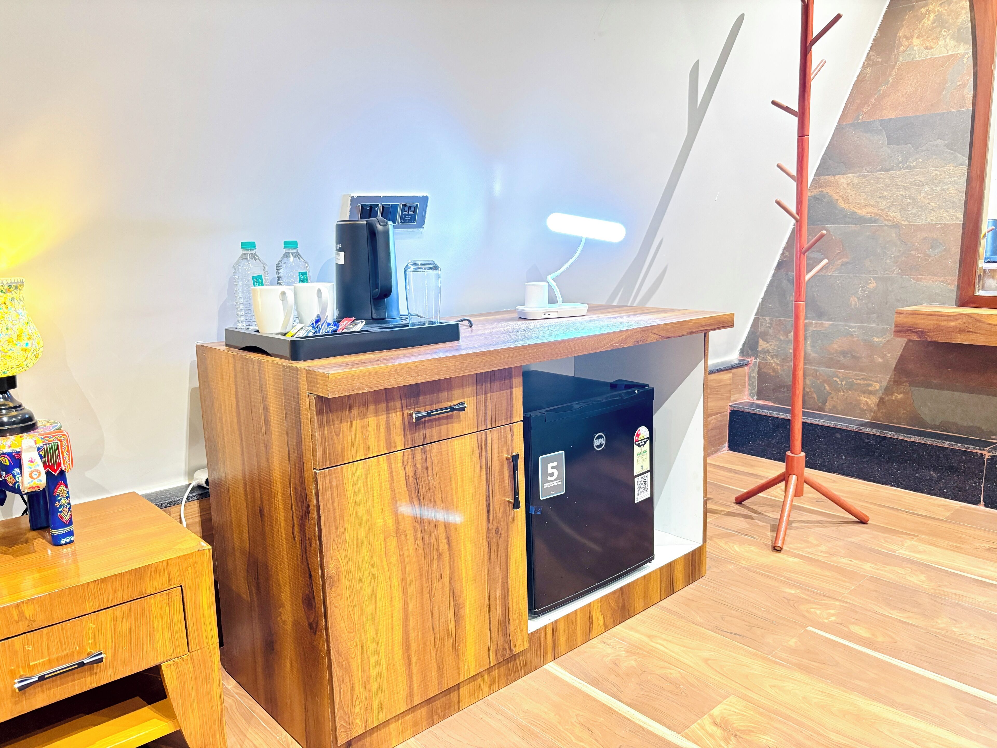 A compact wooden desk with a coffee machine, a glass, and a cup, a small refrigerator, and a wooden nightstand with a lamp.