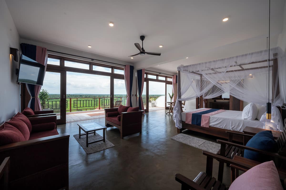 A spacious room with a large bed, a sofa, and a TV. There are large windows and a balcony with a view of the landscape.