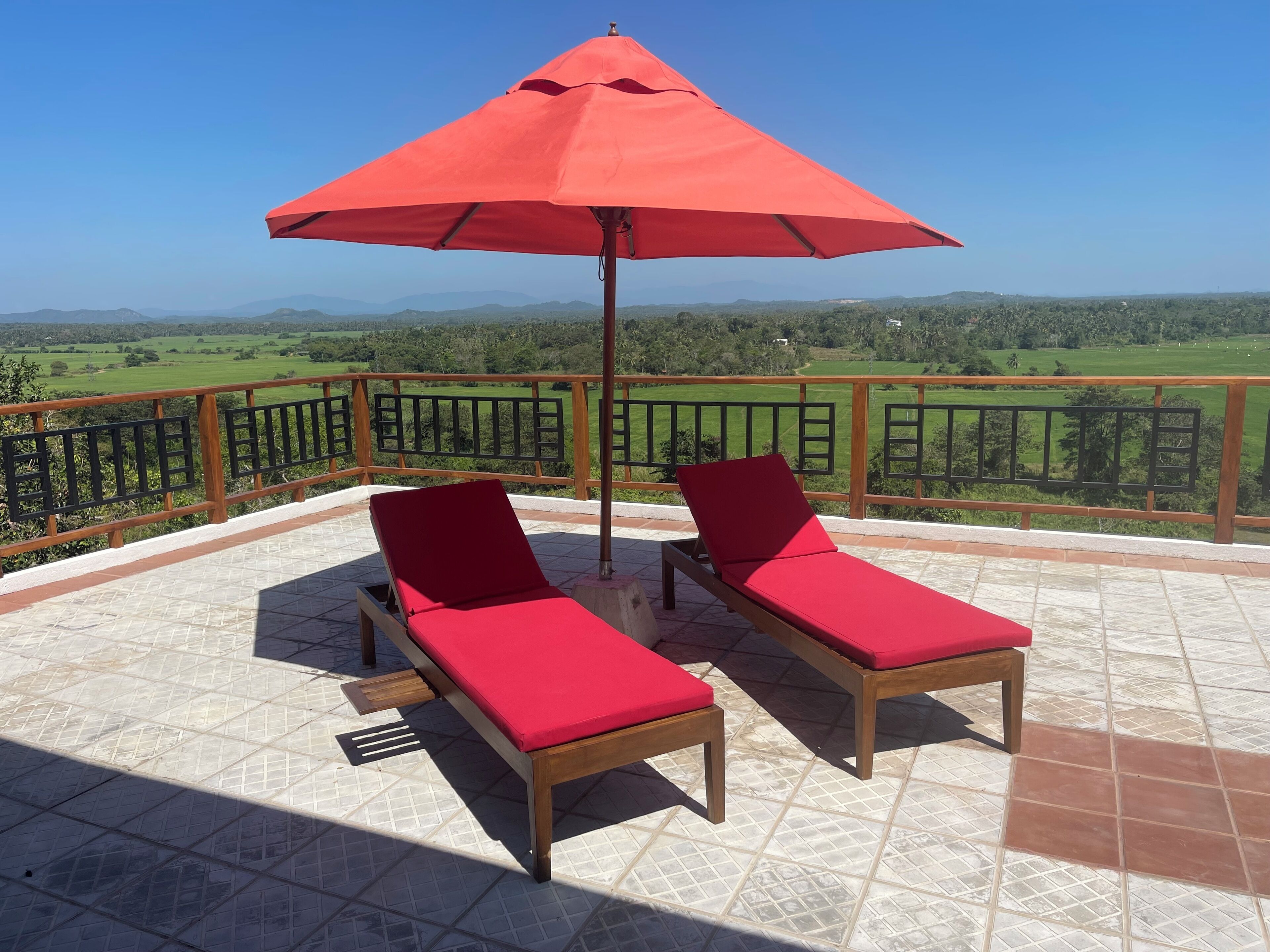 A balcony with two wooden lounge chairs and a red umbrella, overlooking a green landscape.