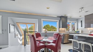 A modern kitchen with a dining area, featuring a glass-top table with red chairs, a stainless steel dishwasher, and a large window with a view of trees. - Top-level duplex featuring a sleek open concept kitchen with stainless appliances, cozy fireplace, stylish furnishings, and in-unit washer/dryer. Designed for basking in the sun under skylights in thoughtful details throughout. (Vancouver)