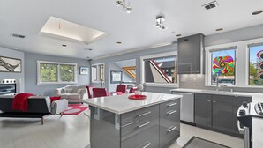 A modern kitchen with a central island, grey cabinetry, and a large window with a colorful view. - Top-level duplex featuring a sleek open concept kitchen with stainless appliances, cozy fireplace, stylish furnishings, and in-unit washer/dryer. Designed for basking in the sun under skylights in thoughtful details throughout. (Vancouver)