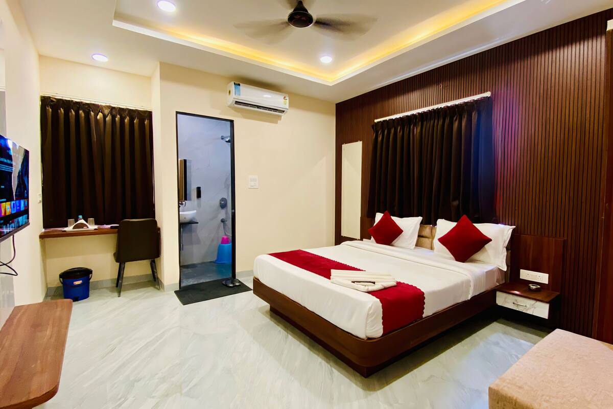 A hotel room with a bed, a TV, a desk, and a bathroom visible through a glass partition.