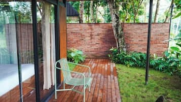 A wooden deck with a metal chair, a glass wall, and a brick wall with greenery.