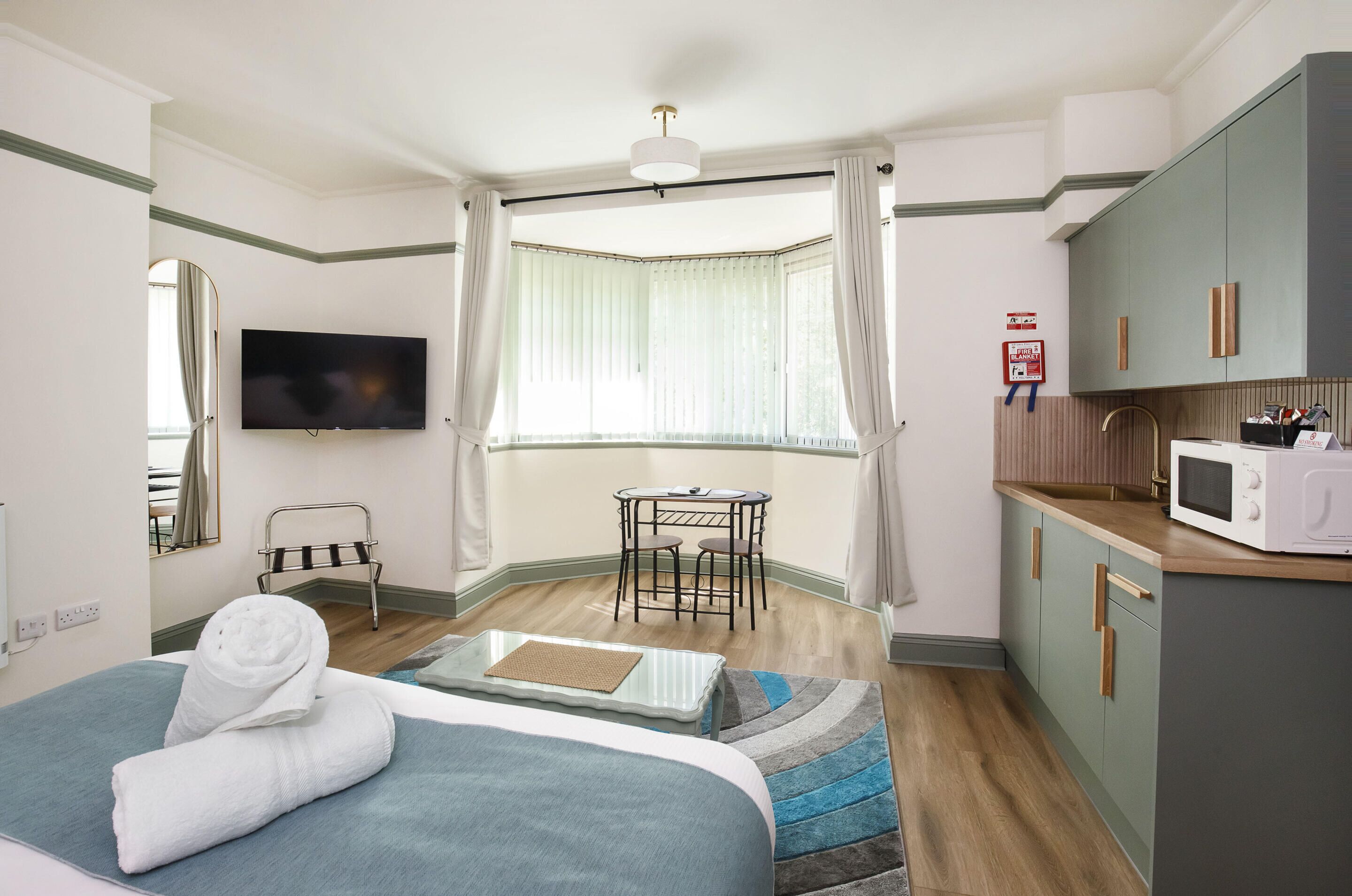 A hotel room with a bed, a TV, a small table with chairs, a window with curtains, a kitchenette, and a fire safety sign.