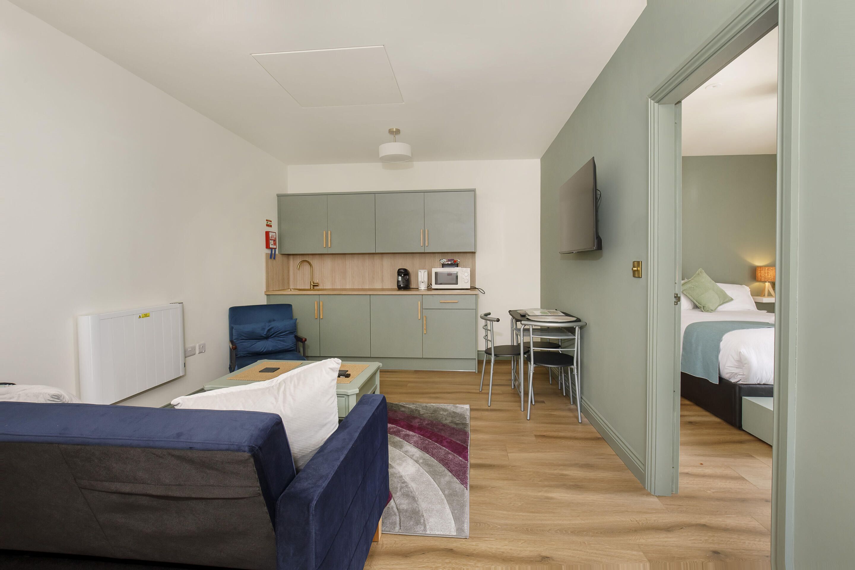 A compact hotel room with a kitchenette, a bed, a seating area with a sofa and armchairs, and a small dining table.