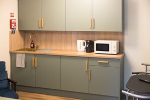 Eine Küchenzeile mit Mikrowelle, Kaffeemaschine und Spüle. An der Wand hängt ein Feuerlöscher und ein Handtuch befindet sich am Schrank.