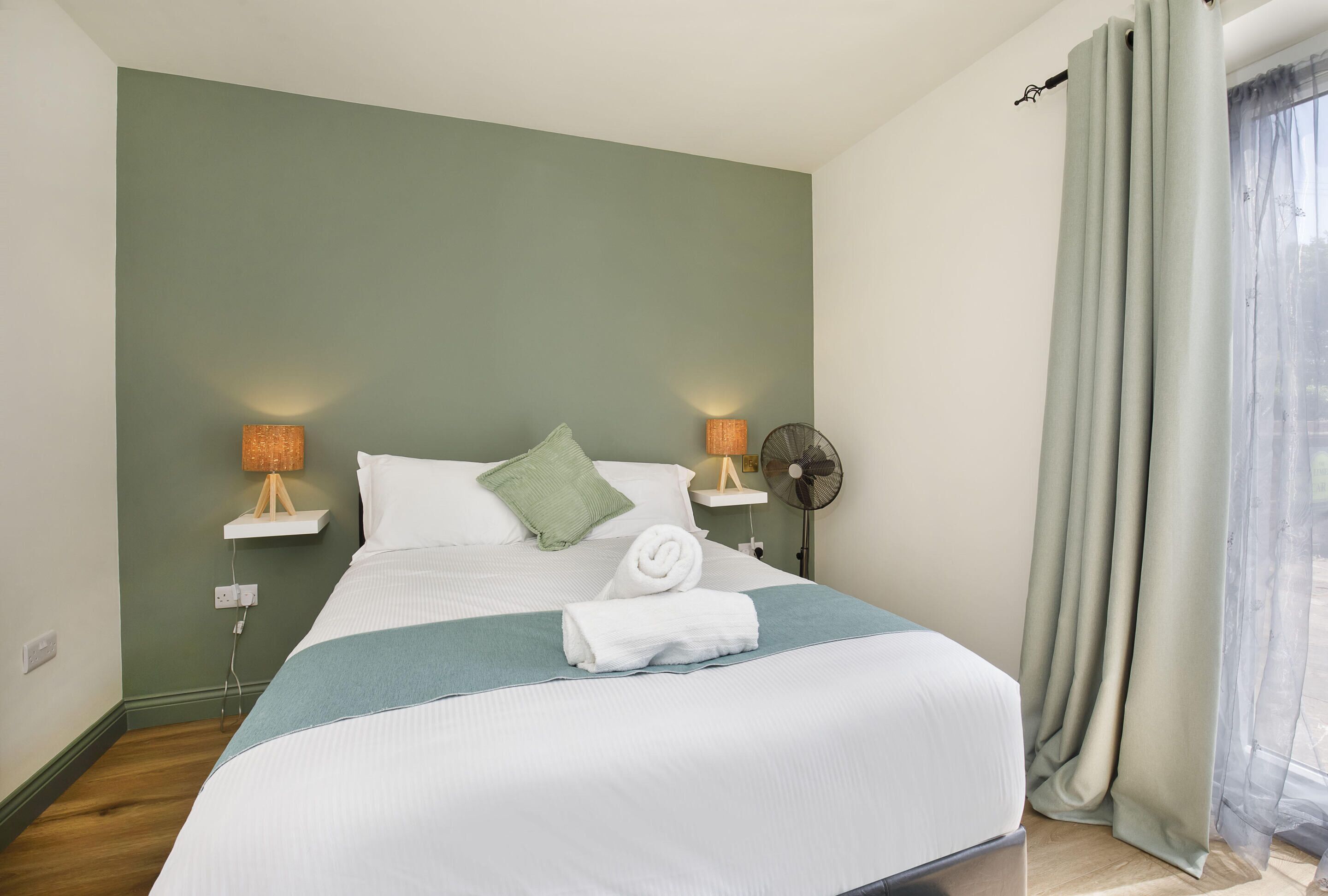 A neatly made bed with a white comforter and a blue throw blanket, two bedside lamps, a wall-mounted fan, and a window with sheer curtains.