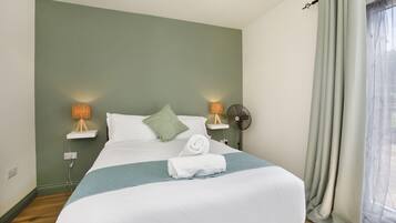 A neatly made bed with a white comforter and a blue throw blanket, two bedside lamps, a wall-mounted fan, and a window with sheer curtains.