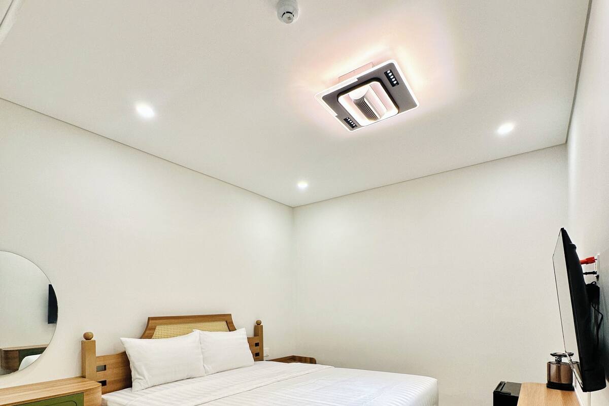 A modern hotel room with a wooden bed, a desk with a chair, a flat-screen TV, and a ceiling-mounted light fixture.