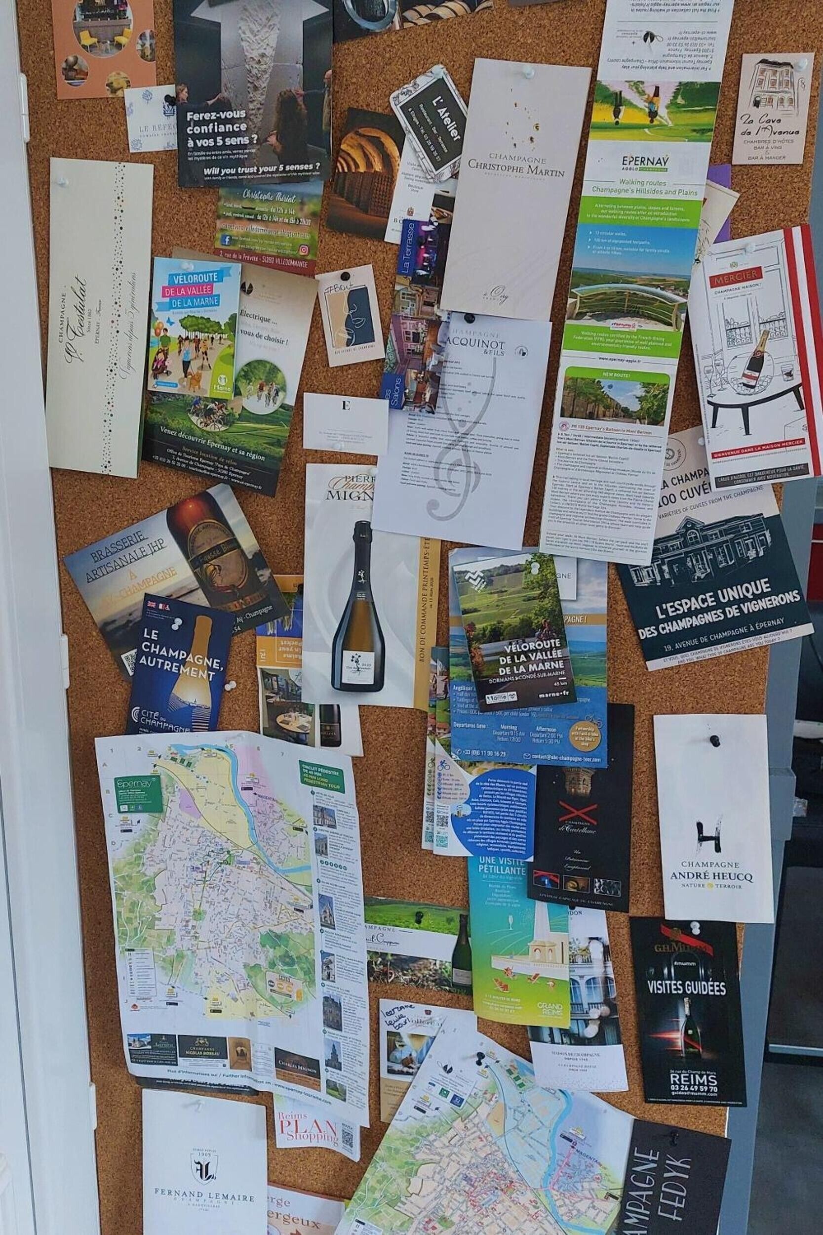 Eine Pinnwand mit verschiedenen Reisebroschüren, Karten und einer angehefteten Weinflasche.