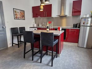 A modern kitchen with red cabinets, a stainless steel refrigerator, a dining table with chairs, and a pendant light.