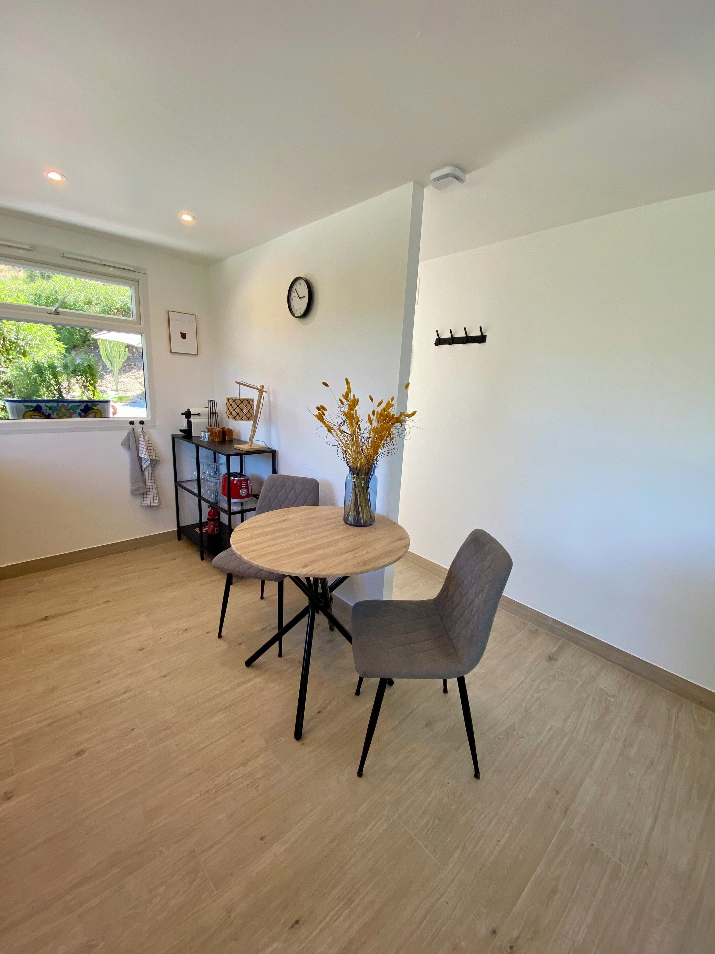 A modern dining area with a round wooden table and two grey chairs, a vase with dried flowers, a clock on the wall, and a window with a view of greenery.
