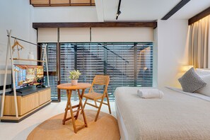 Une chambre d’hôtel moderne avec un grand lit, une petite table ronde avec une chaise, une plante en pot et une vue sur le paysage urbain à travers de grandes fenêtres du sol au plafond.