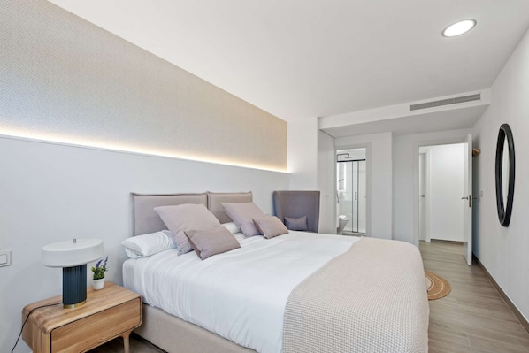 A modern bedroom with a large bed, bedside table with a lamp, and a wall-mounted mirror.