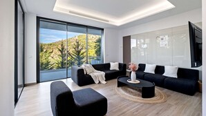 A modern living room with a sectional sofa, a coffee table, and a large window offering a view of a forested hillside. - Villa Pinares de San Antón Sea, City & Mountain Views (Málaga)