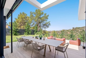 A modern outdoor dining area with a long table and chairs, surrounded by a wooden deck and overlooking a scenic landscape with trees and hills.
