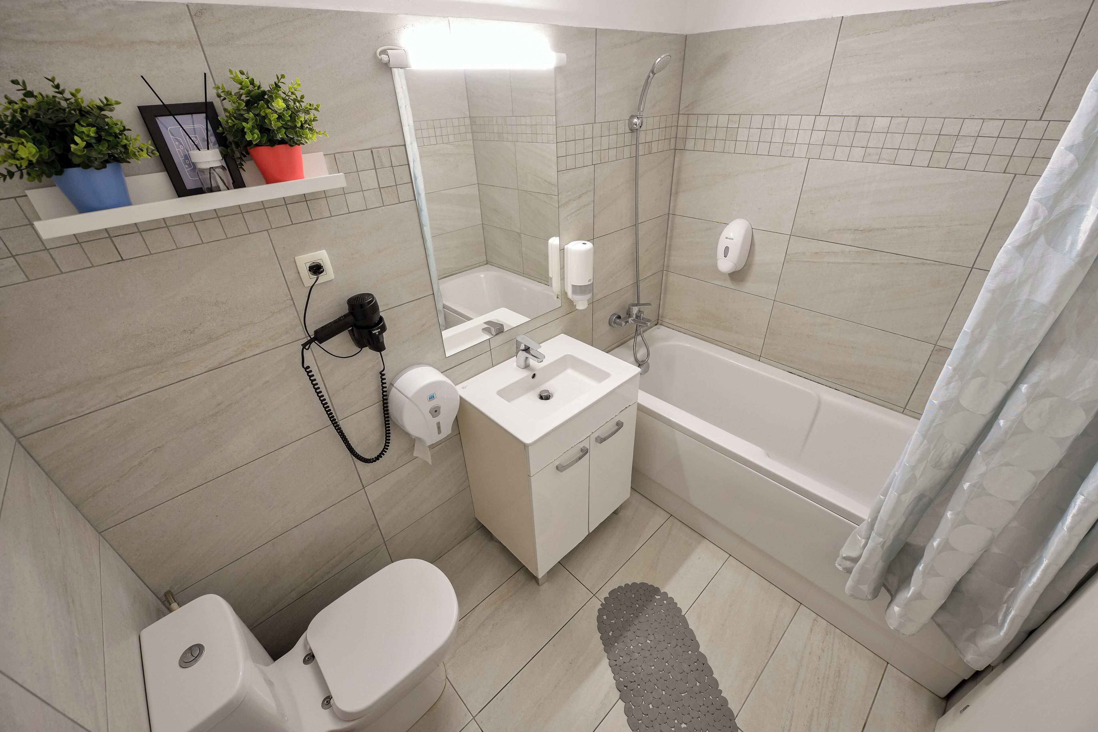 Un baño moderno con inodoro, lavamanos y ducha. Hay plantas en macetas sobre una repisa, un perchero y un espejo.